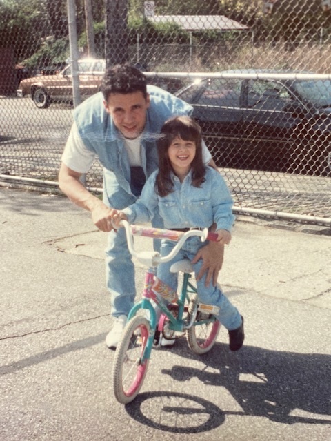 Jaina riding a bicycle as a little girl and a man smiling at the camera.